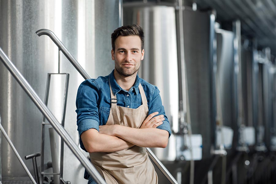 Specialized Business Insurance - Brewer Wearing Tan Apron Leans Against Large Tanks in His Brewery
